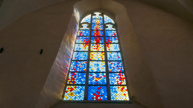 Tall pointed stained-glass window centered within a deep plaster reveal, filled with blue, red and yellow geometric panes.