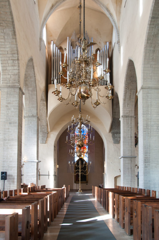 Central aisle of St Nicholas Church beneath a large brass chandelier, with pointed arcades, wooden seating, and a colorful suspended installation.