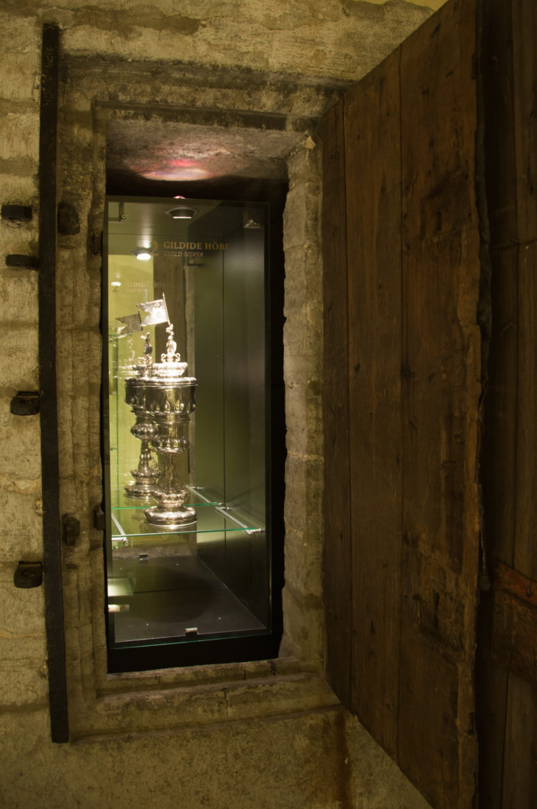 Illuminated silver vessel in a glass case inside a low stone chamber with heavy timbers and an open plank door.