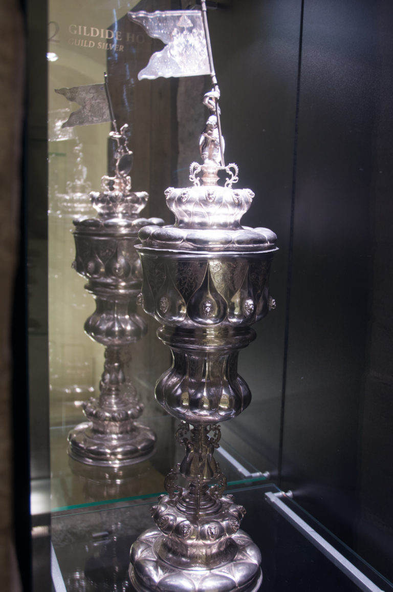 Close view of ornate silver vessels with stems, lids, finials, and reflected surfaces inside a museum display case.