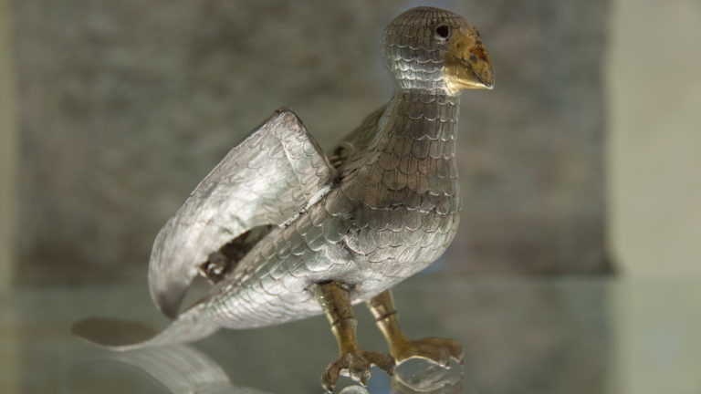 Small silver bird-form object standing on two legs with raised wings, scaled feather decoration, and a warm-toned beak.