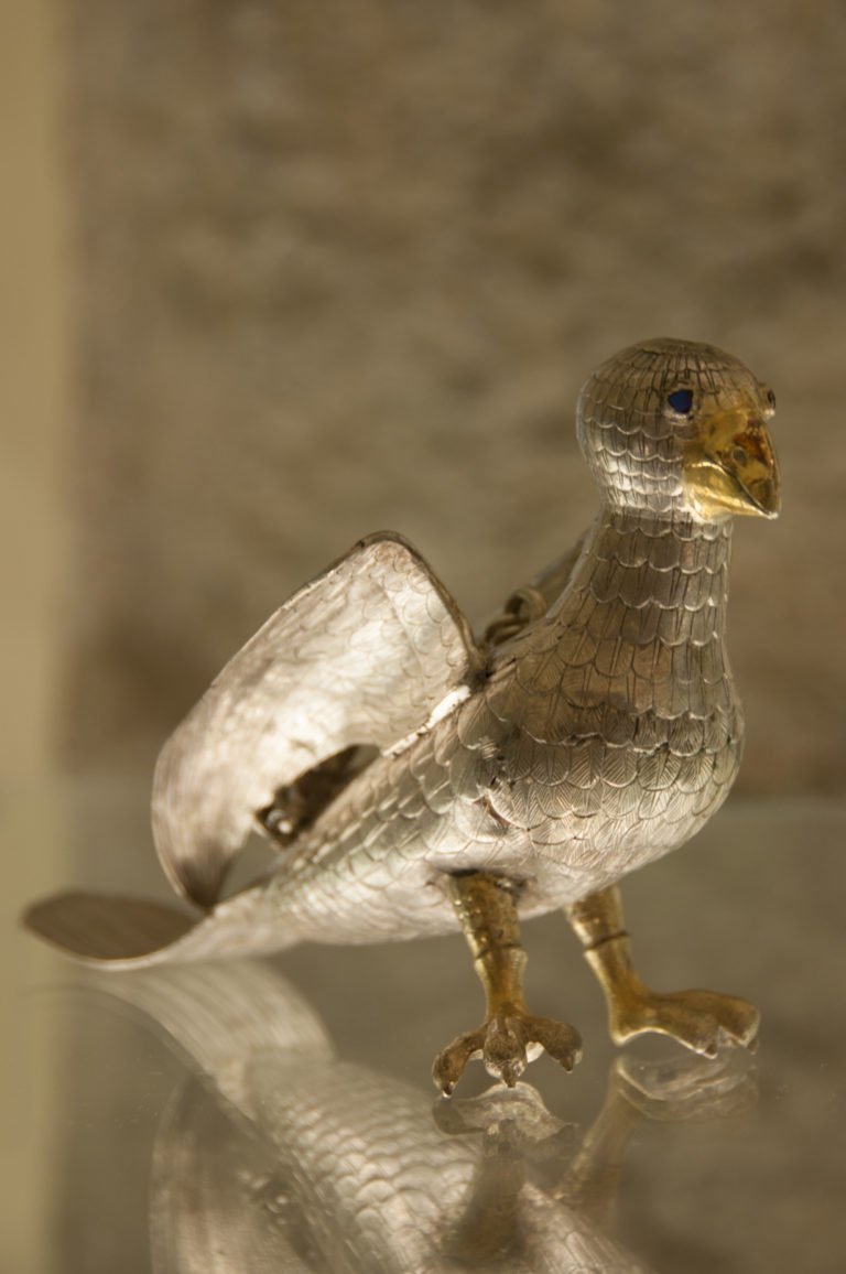 Side view of a silver bird-form object with raised wings, patterned plumage, slender legs, and a warm-toned beak.