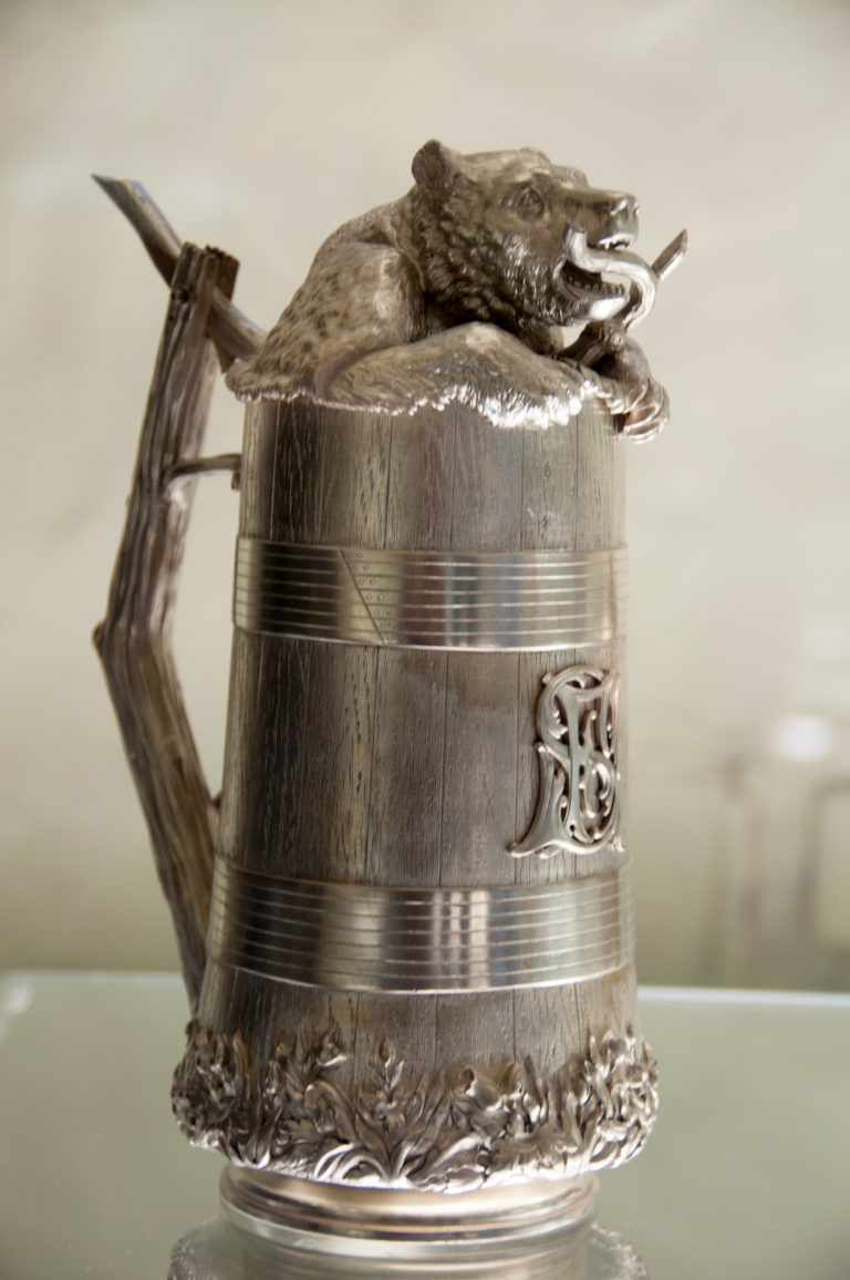 Ornate cylindrical silver tankard with horizontal bands, raised monogram, decorated foot, and sculptural handle and rim fittings.