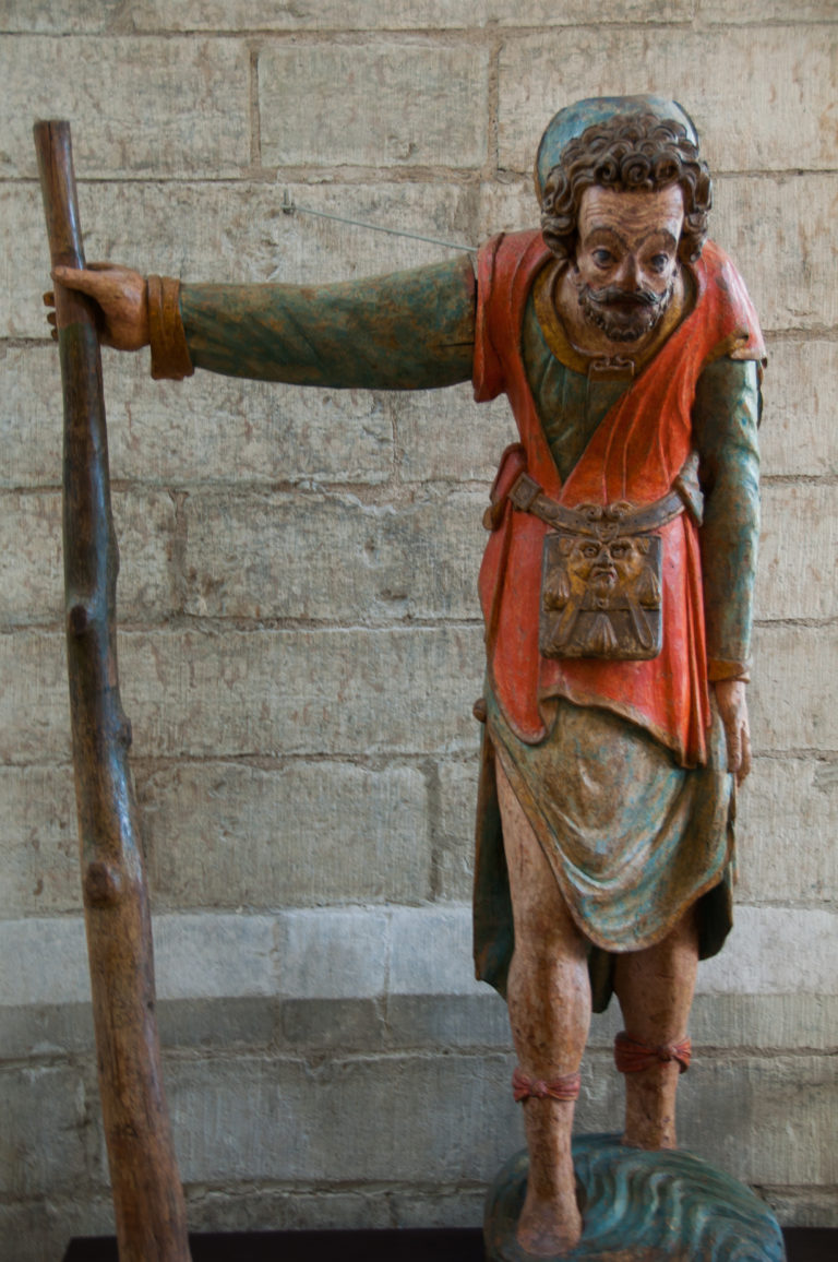 Polychrome carved wooden man leaning forward while holding a long vertical staff, wearing red and green garments and a waist pouch.