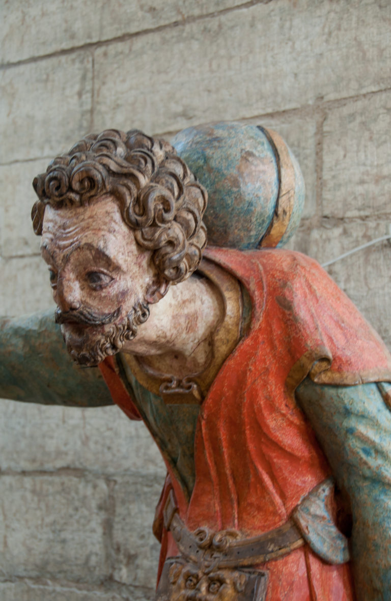 Close view of a bearded polychrome wooden figure with curled hair, red outer garment, and blue-green sleeve.