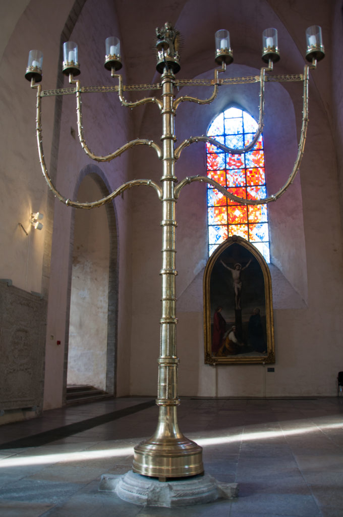 Tall freestanding metal candelabrum with curved arms and multiple candle sockets, silhouetted before a colored church window.