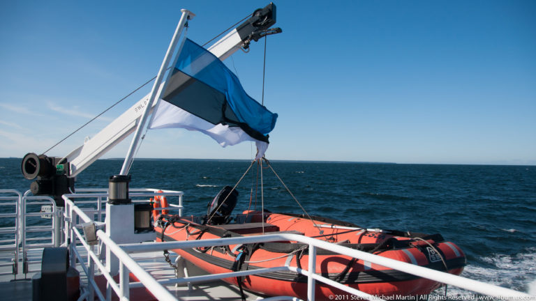 Estonian flag flying above orange rescue craft, white railings, and a deck crane on a passenger ferry at sea.