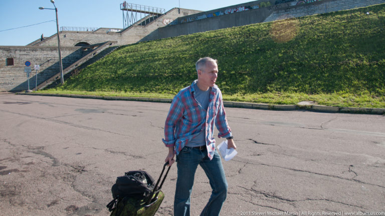 Traveler pulling wheeled luggage beside the grassy concrete terraces and stairs of Tallinn Linnahall.