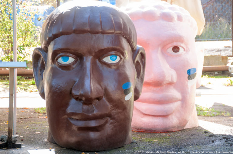 Two oversized sculpted human heads outdoors, one dark brown with blue eyes and one pale pink behind it.