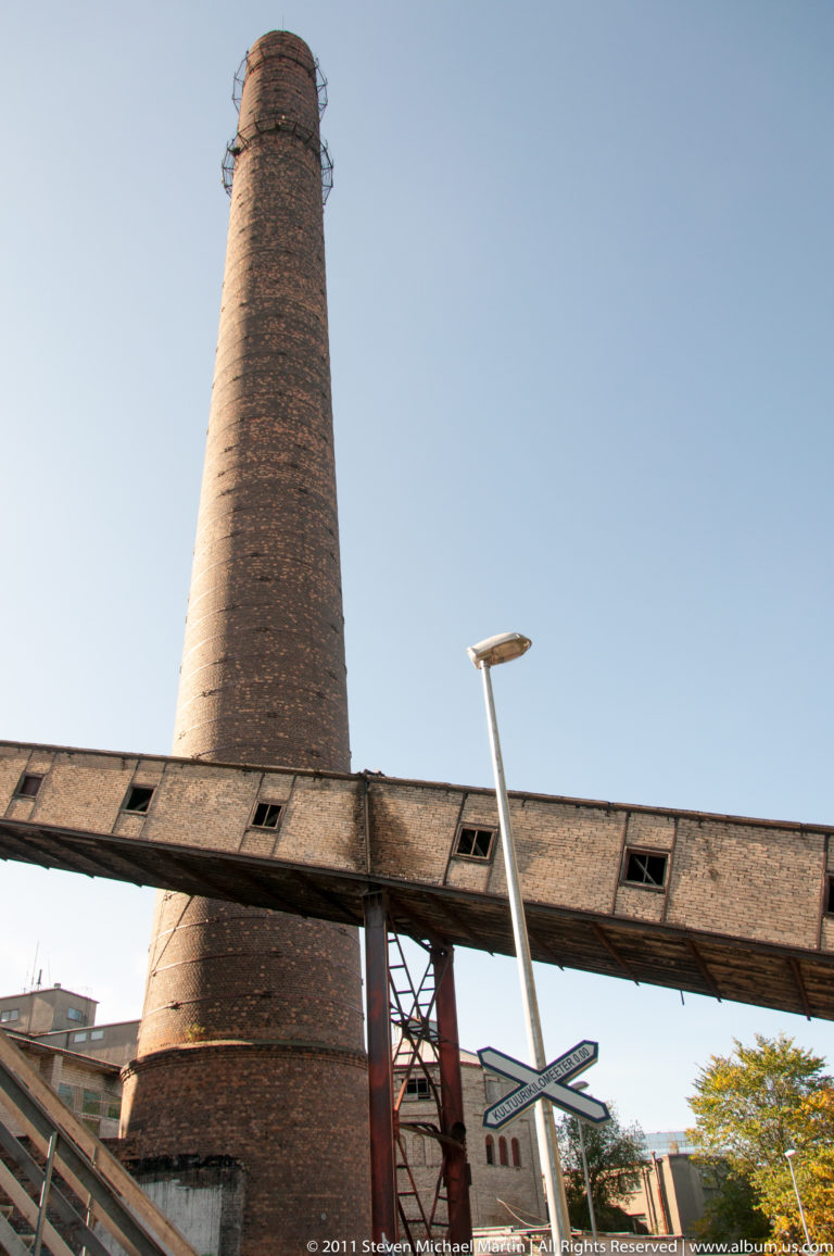 Tall tapered brick industrial chimney rising behind an elevated metal-and-roofed structure beneath a clear blue sky.