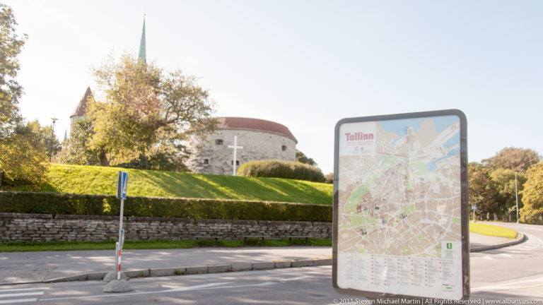 Visitor map beside a Tallinn roadway with a massive round medieval stone tower and red roof rising behind trees.