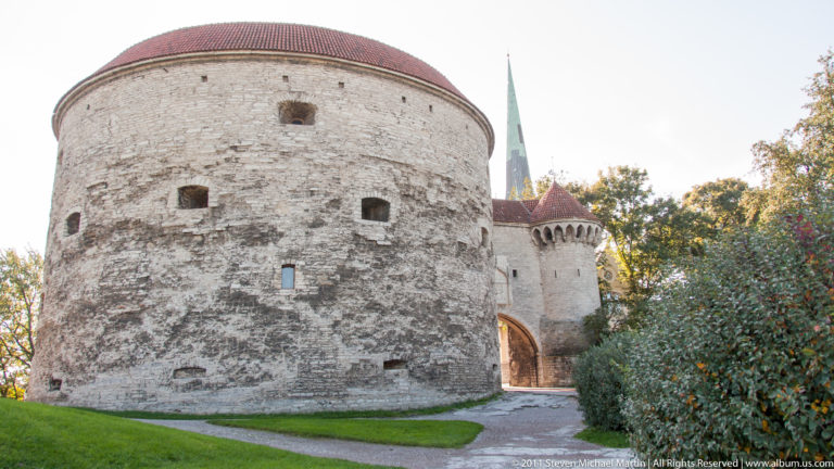 Massive round Fat Margaret tower with small defensive openings, red roof, adjoining gateway, and green church spire behind.