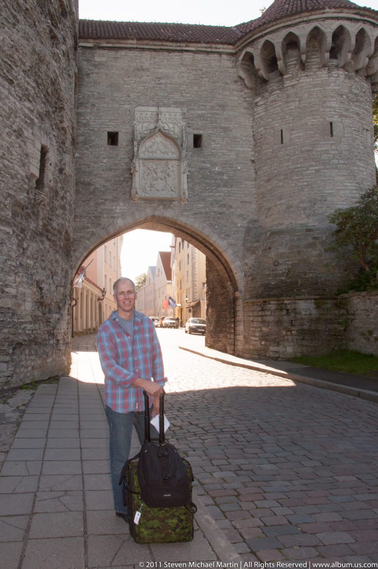 Adult with rolling luggage standing on cobblestones before the broad stone arch of Tallinn’s Great Coastal Gate.