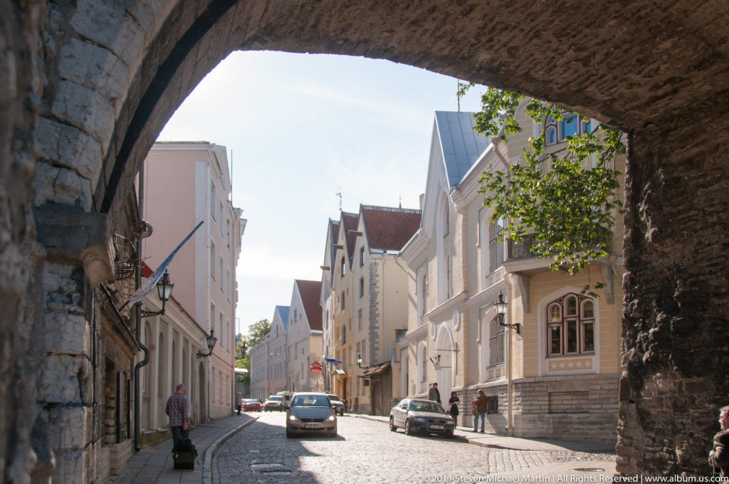 Sunlit Tallinn street with historic buildings and parked cars framed through a dark medieval stone gateway arch.
