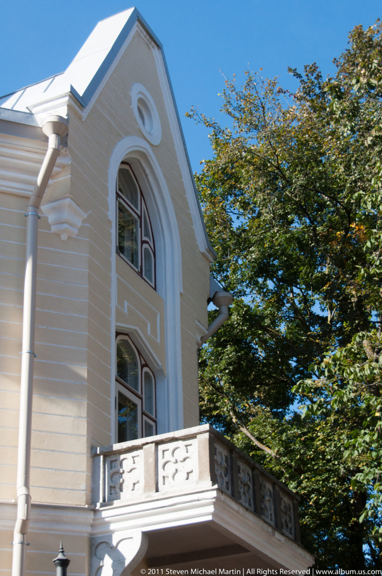 Pale gabled historic house with tall arched windows, projecting white balcony, circular gable ornament, and trees.