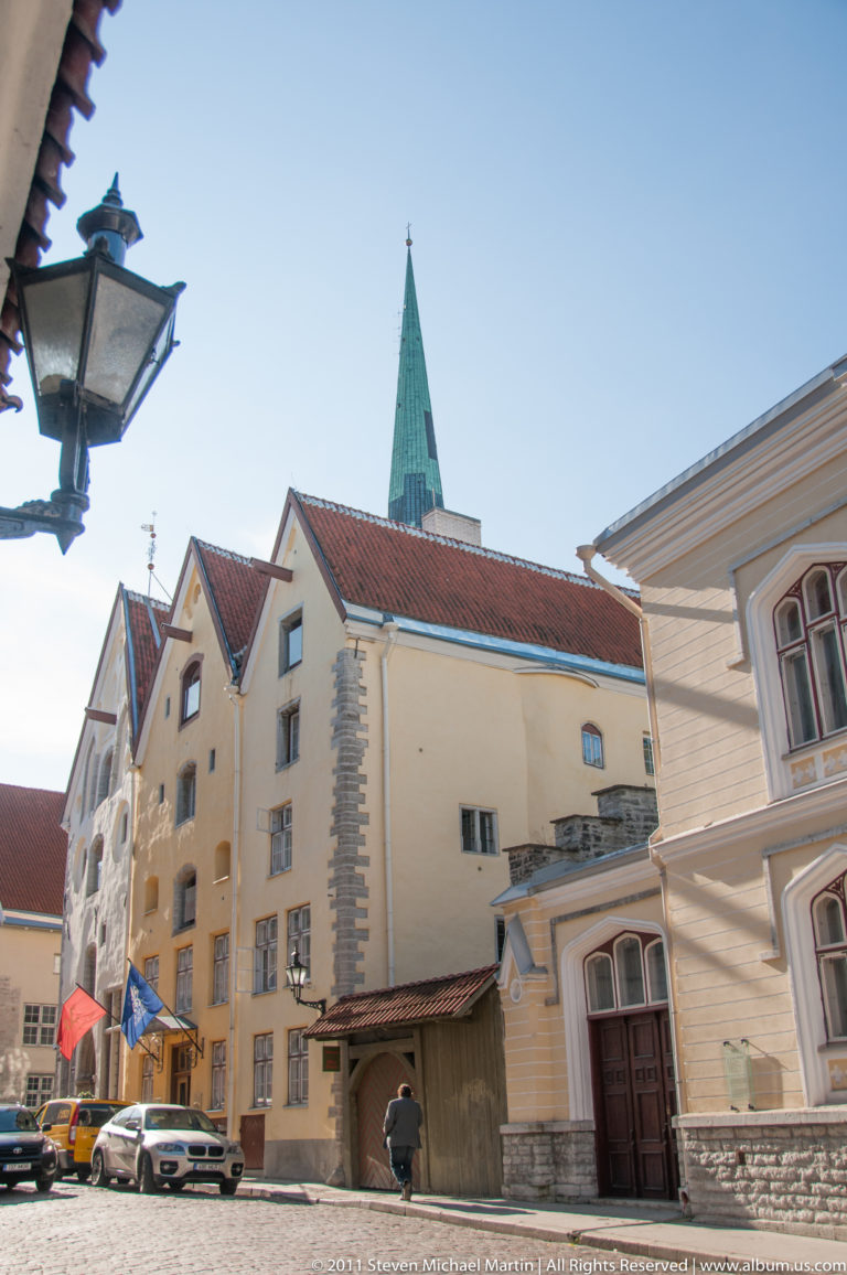 Quiet Tallinn courtyard with pale gabled historic buildings, red roofs, a timber structure, and a green church spire beyond.