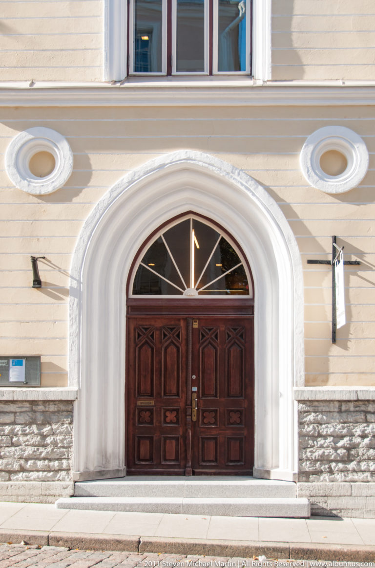 Dark wooden double doors beneath a fanlight in a broad white arch, flanked by oval decorative medallions.