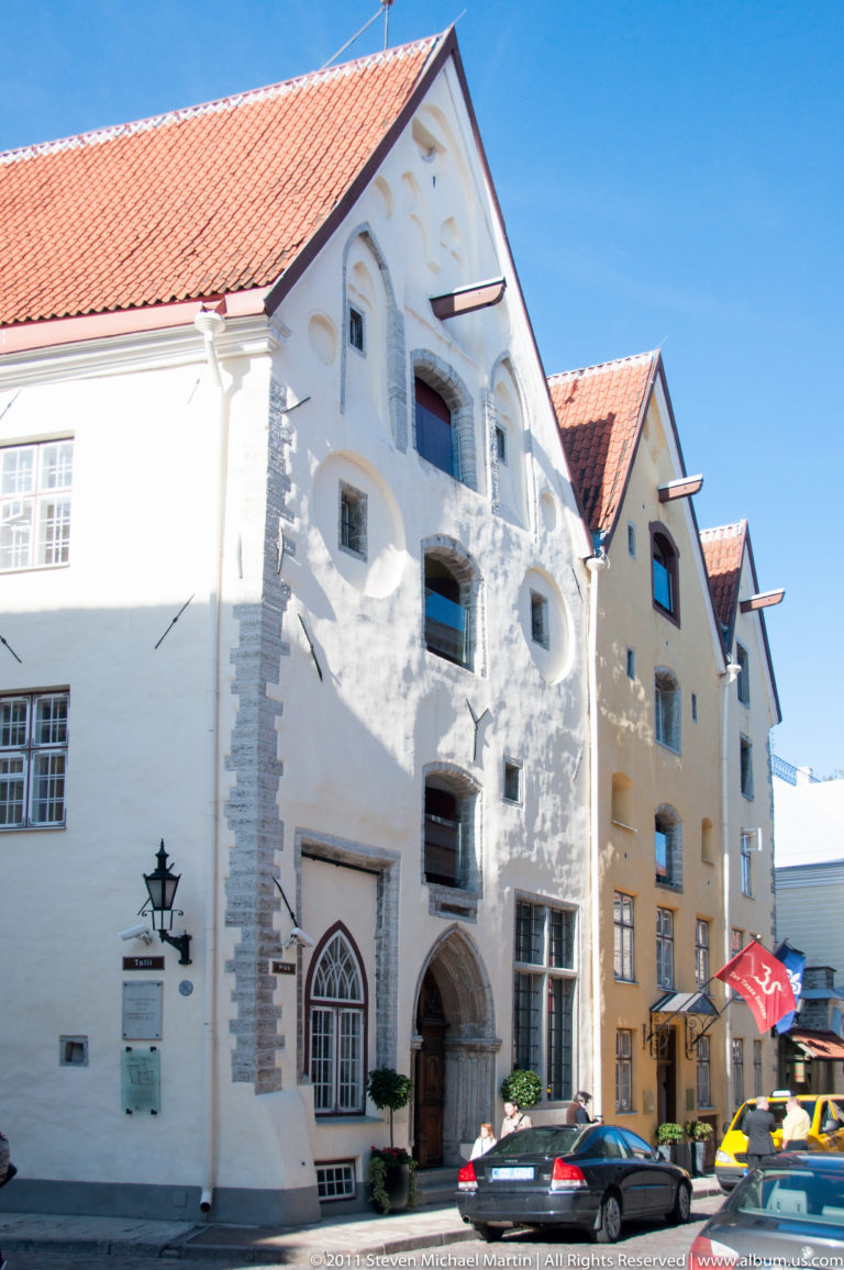 White gabled historic building with irregular small windows, arched doorway, thick masonry, and red-tiled roof.