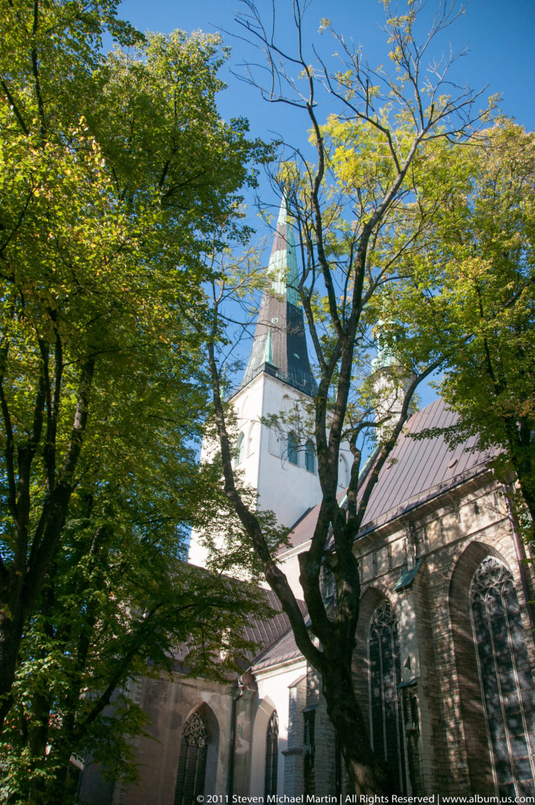 Stone church with tall Gothic windows and a steep green spire partly obscured by leafy trees.