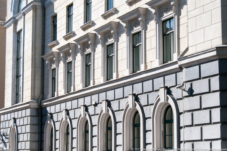 Pale classical stone façade with arched lower windows, tall upper windows, projecting cornices, and sculpted surrounds.