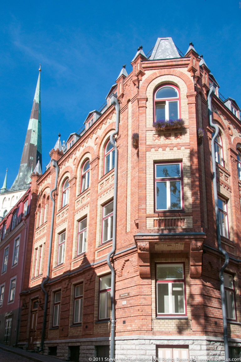 Ornate red-brick corner building with arched windows, decorative gables, and a green church spire behind.