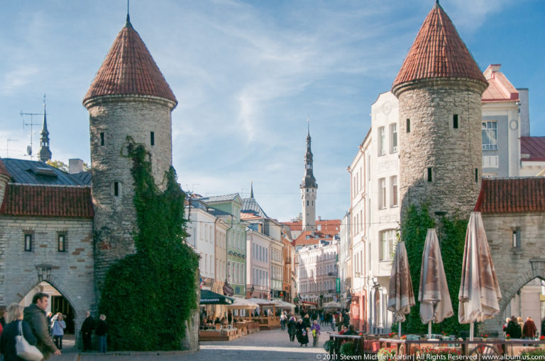 Viru Gate towers framing a busy Tallinn Old Town street with ivy, pedestrians, cafés, and the Town Hall tower in the distance.