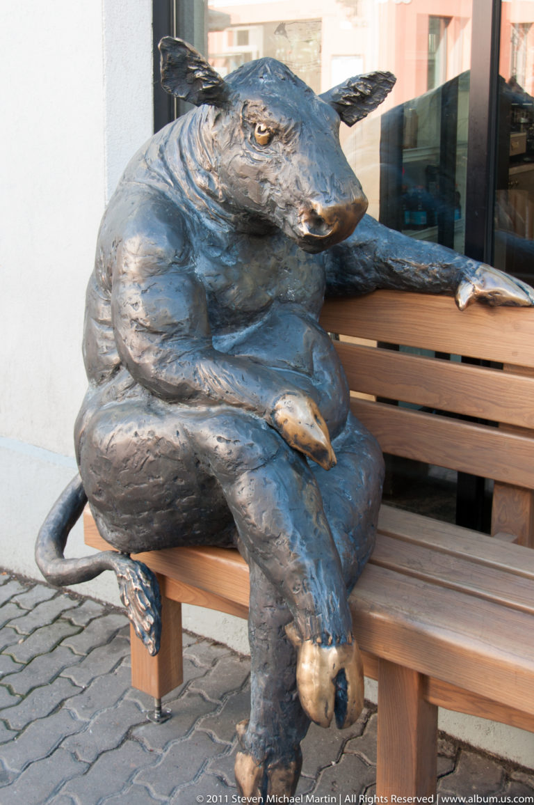 Bronze sculpture of several animal figures seated together on a wooden bench beside a public walkway.