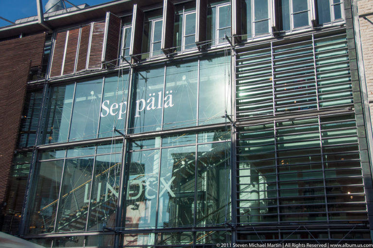 Modern glass façade with horizontal metal louvers, structural framing, and large white lettering across the windows.