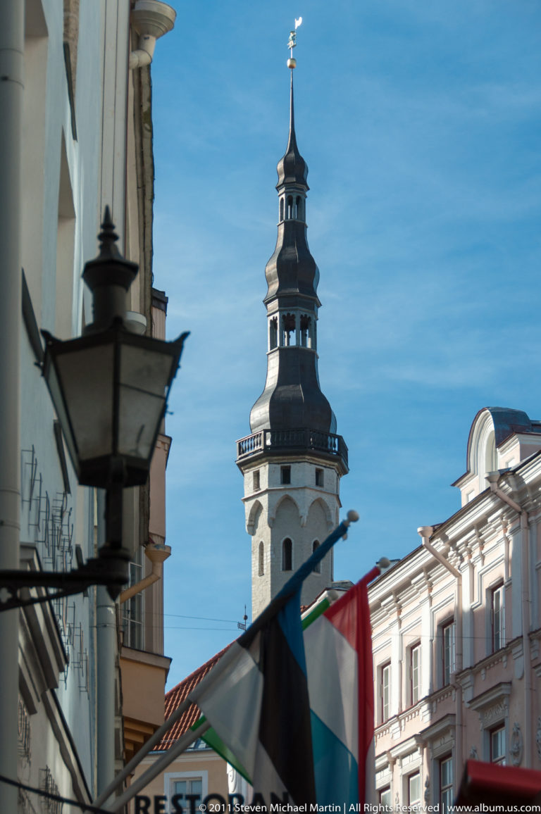 Tallinn Town Hall tower rising above rooftops, framed by a black street lantern and restaurant awnings.