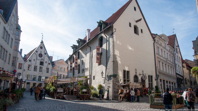 Cobbled Tallinn Old Town street lined with steep-gabled buildings, restaurant terraces, signs, and pedestrians.