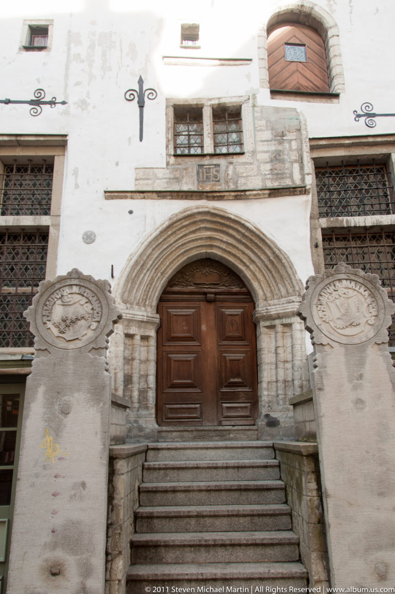Pointed stone portal with heavy wooden double doors, steps, carved details, and circular stone plaques on a historic façade.