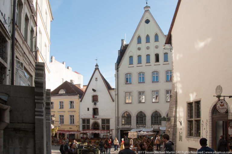 Steep-gabled historic buildings overlooking market stalls and pedestrians in Tallinn Old Town under a clear blue sky.