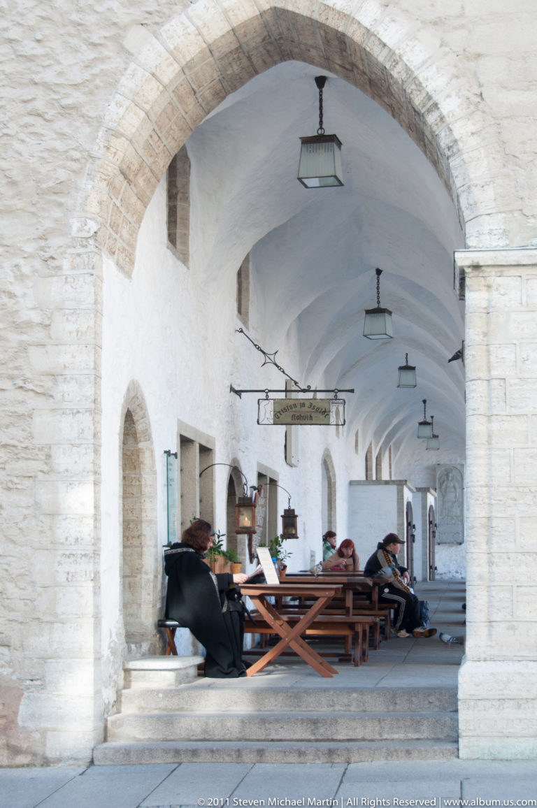 Pointed stone arch opening into Tallinn Town Hall’s covered arcade with café tables, chairs, and hanging lamps.