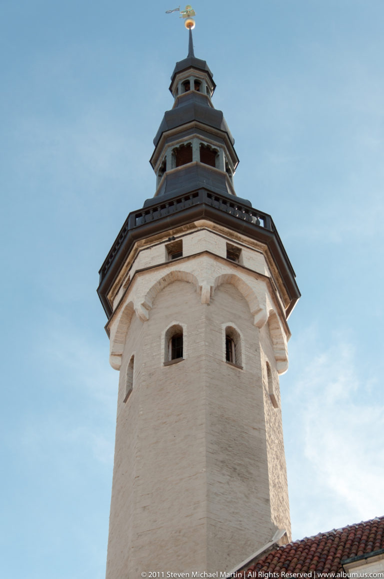 Upper Tallinn Town Hall tower with pale masonry shaft, dark gallery stages, tiered spire, and small arched openings.