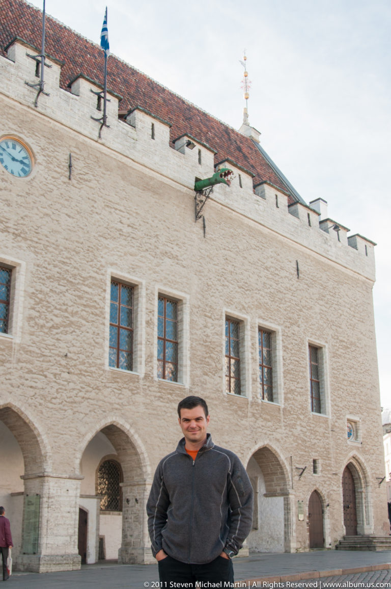 Traveler standing before Tallinn Town Hall’s pale limestone façade, pointed arcades, rectangular windows, and red-tiled roof.