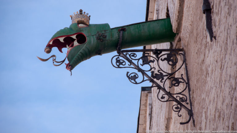 Green dragon-shaped rainwater spout with open red mouth projecting from Tallinn Town Hall on ornate iron brackets.