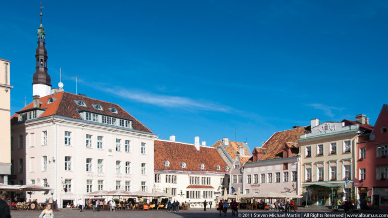 Colorful historic buildings and café seating on Tallinn Town Hall Square with the Town Hall tower rising behind the rooftops.