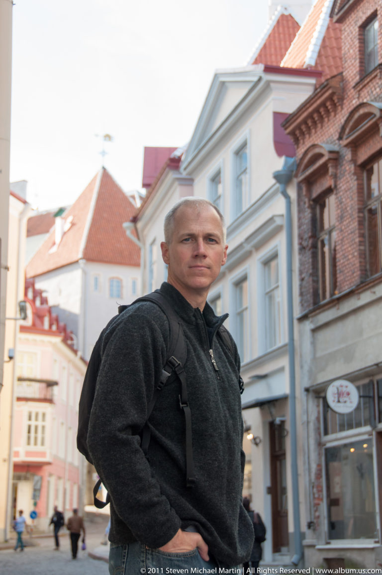 Adult standing on a Tallinn Old Town street with pale and red-brick historic buildings receding behind.