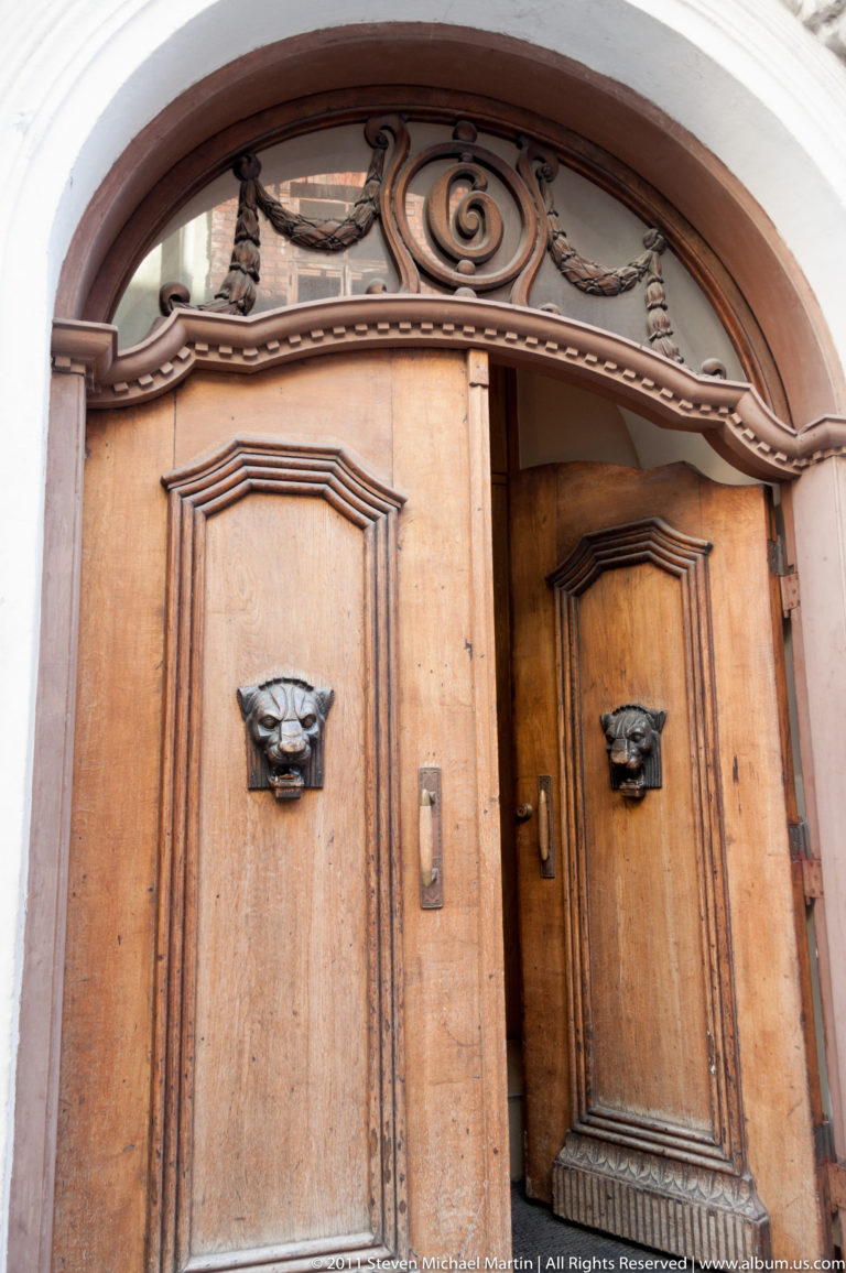 Ornate wooden double doors in a rounded opening, with carved panels, scrollwork, and projecting grotesque face fittings.