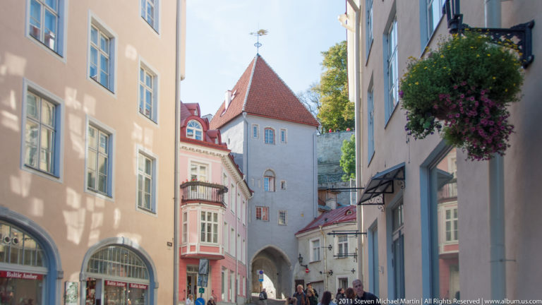 Narrow Tallinn Old Town street leading toward a white medieval gate tower with an arched passage and steep red roof.