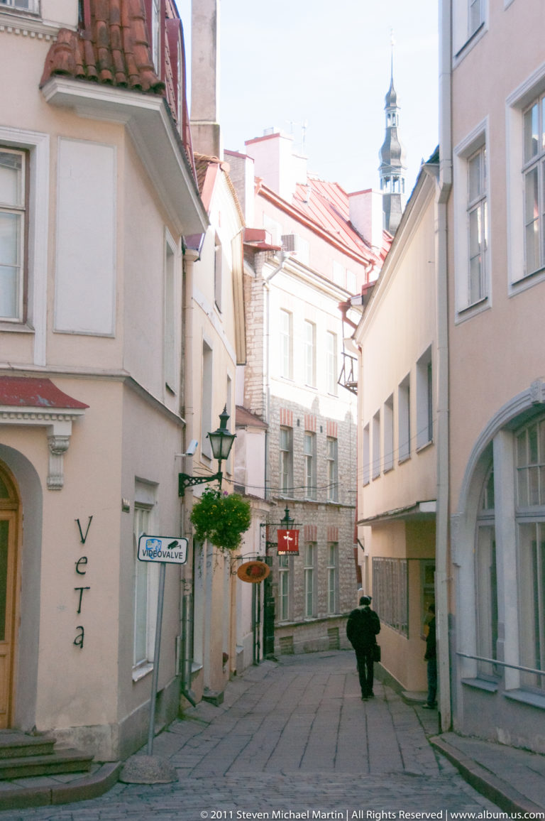 Quiet narrow Tallinn lane with pale historic façades, hanging signs, a pedestrian, and a distant tower above the roofs.