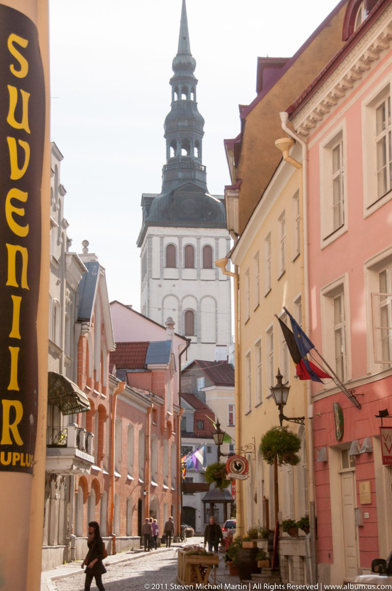 Narrow Tallinn Old Town street with pastel façades, shops, pedestrians, flags, and a tall white church tower in the distance.