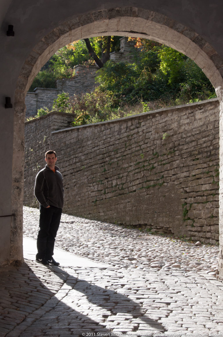 Adult standing beyond a large stone arch on a sunlit cobbled passage beside a high masonry wall.