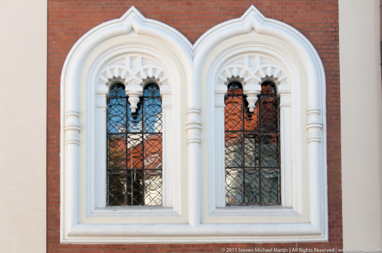 Pair of pointed Gothic windows with white molded surrounds and dark leaded glazing set into a red-brick wall.