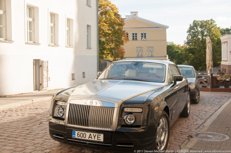Dark Rolls-Royce Phantom parked on a cobblestone Tallinn street between pale historic buildings.