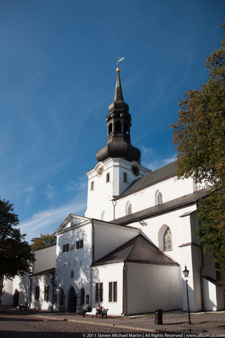 White historic church complex with a dark multi-stage tower and tapering spire under a clear blue sky.