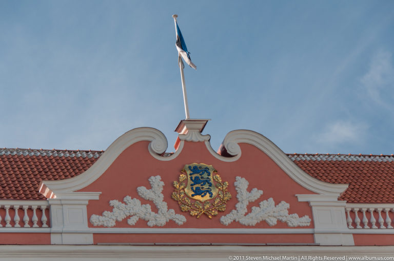 Curved pink gable with Estonia’s three-lions coat of arms, white relief ornament, and flagpole.