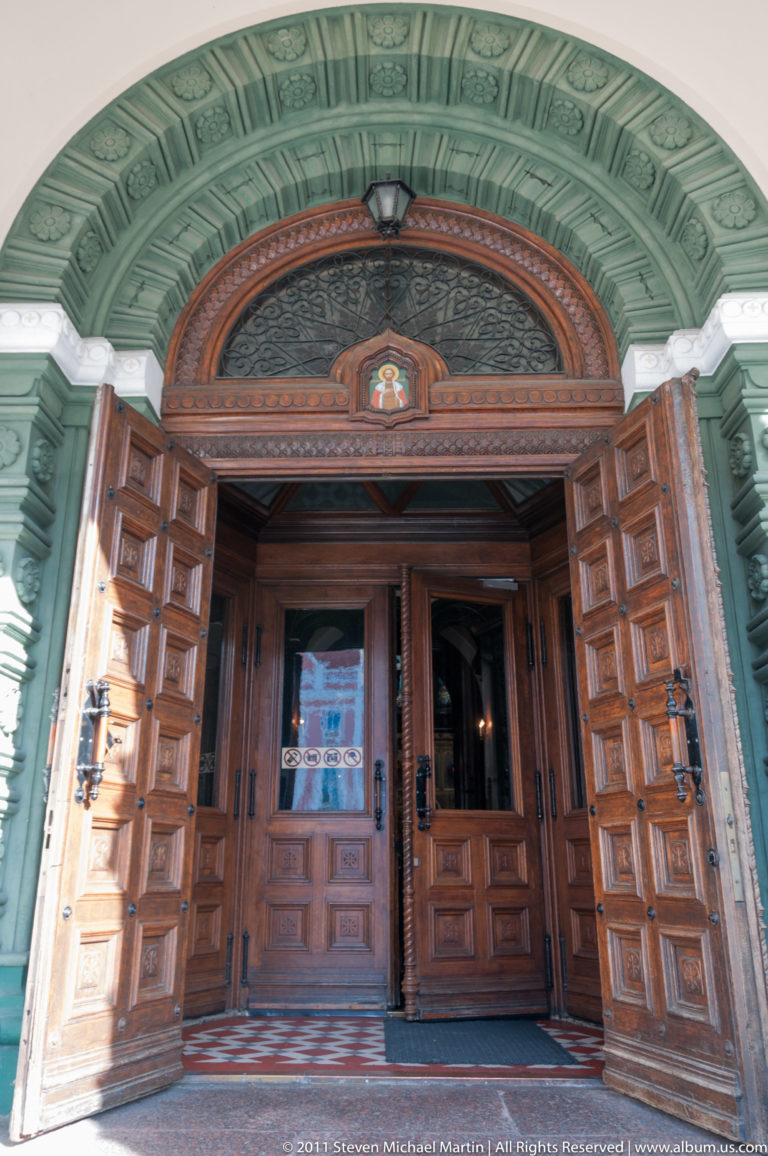 Open paneled wooden doors beneath an ornate green-and-terracotta arched entrance with carved moldings and semicircular transom.