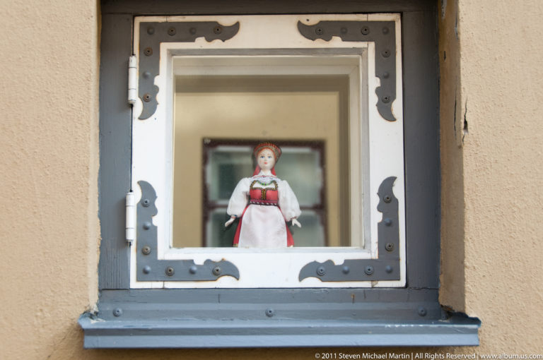 Small doll in traditional-style dress displayed behind a deep-set window with riveted metal corner fittings.