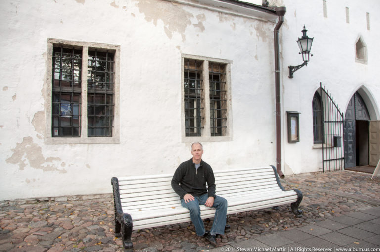 Adult seated on a white bench beside barred windows, a lantern, pointed-arch doors, and cobblestones in Tallinn Old Town.